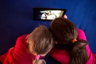 Two young children watching a video on a tablet together, seated on a blue surface.