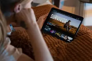 Person watching a streaming platform on a tablet, displaying the show 'Survivor,' while relaxing on a cozy blanket.